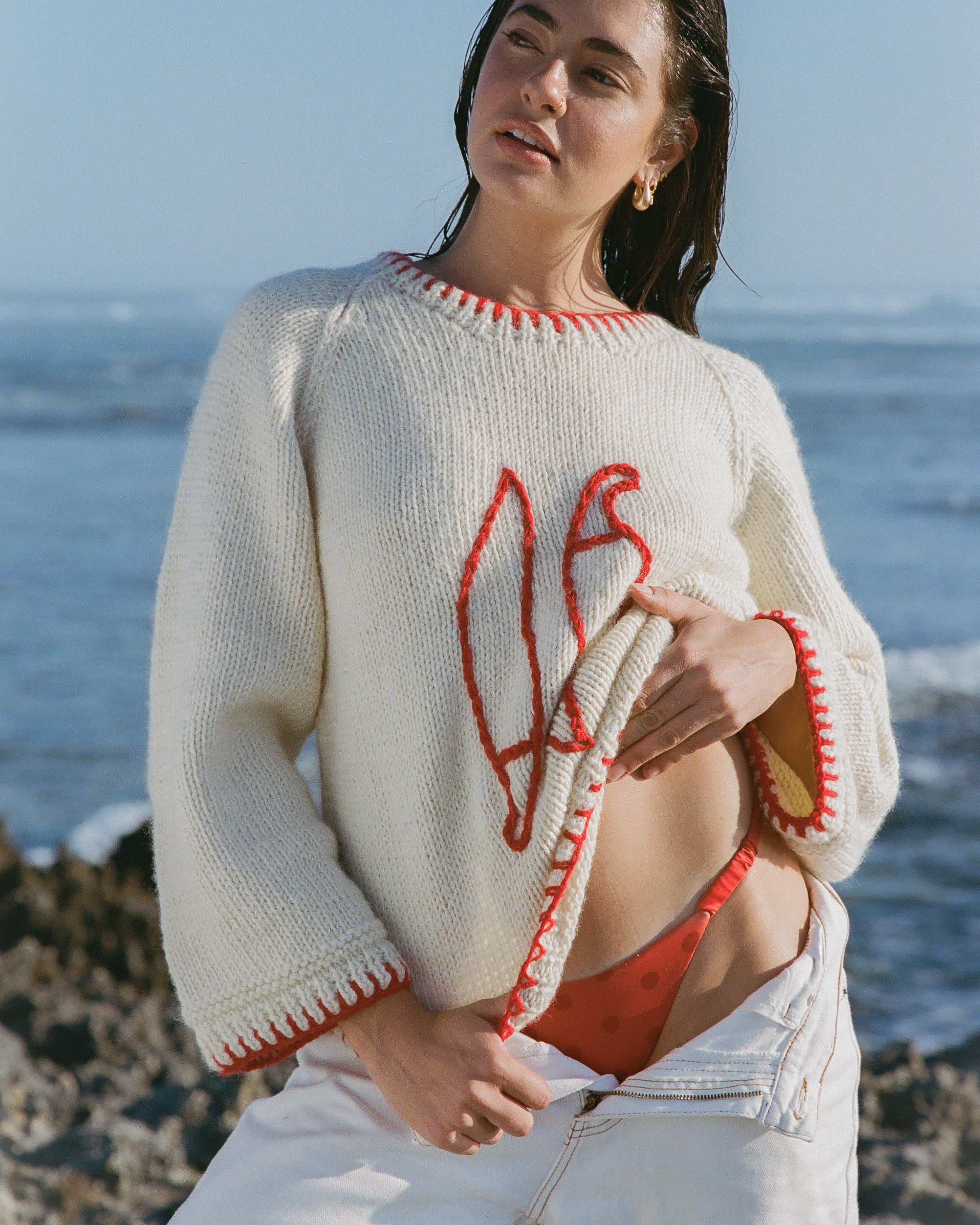 Woman wearing a white sweater with red accents and red bikini standing on a rocky beach.