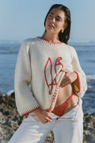 Woman wearing a white sweater with red accents and red bikini standing on a rocky beach.