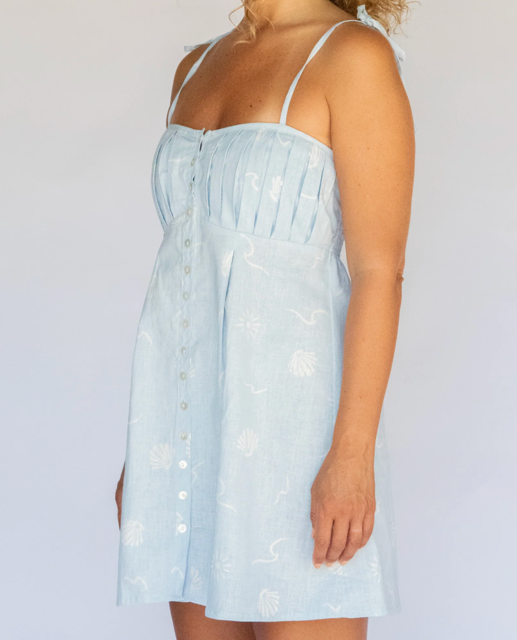 A woman wearing a light blue sleeveless mini dress with thin straps, a gathered bust, front buttons, and a subtle tonal floral pattern, photographed against a plain light background.